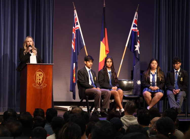 Principal and students on stage in the school Hall with flags behind them